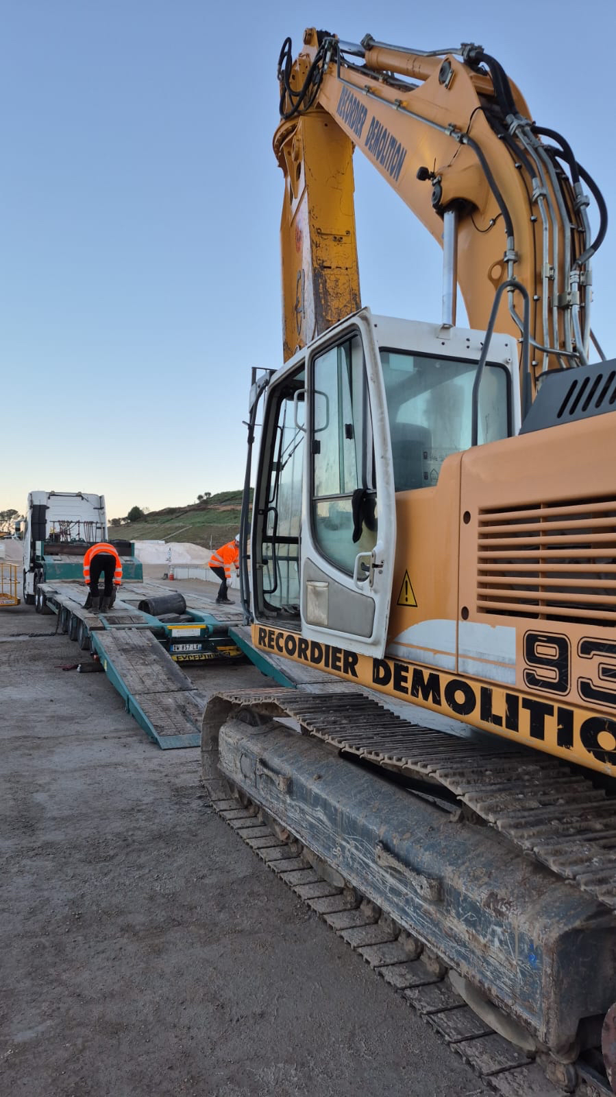Une pelleteuse jaune sur une remorque, des ouvriers en gilets orange se préparent au transport sur un chantier de construction.