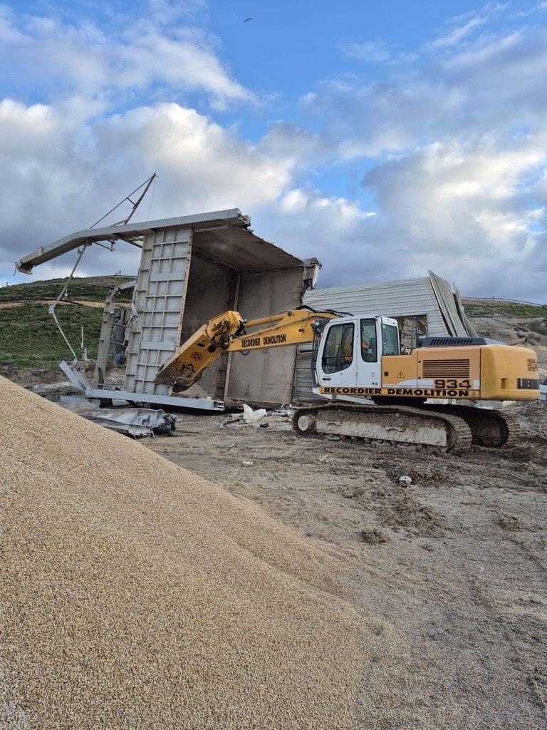 Une pelleteuse jaune démolit une structure en béton à l'extérieur, sous un ciel nuageux.