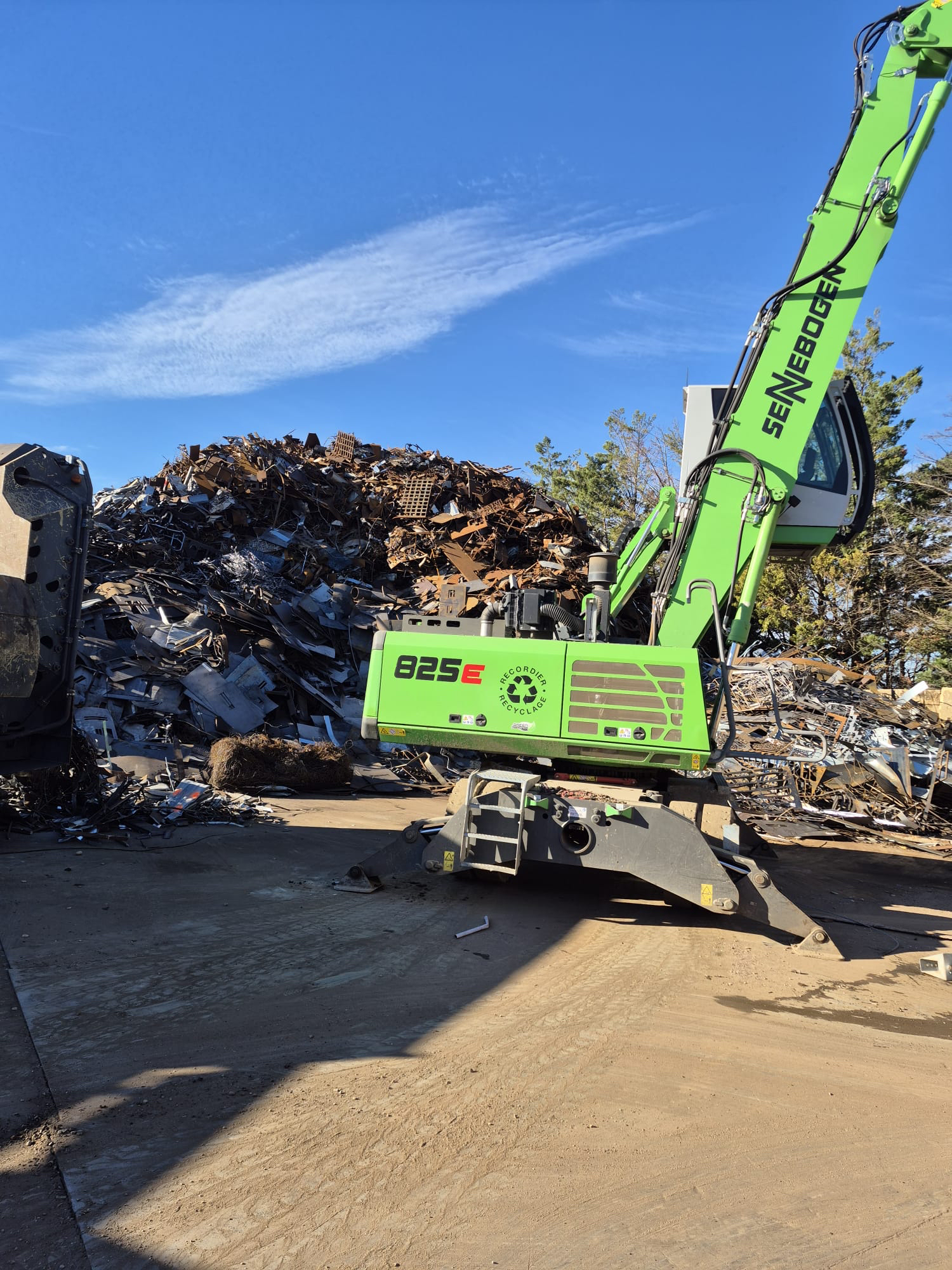 Un engin de manutention vert trie des déchets métalliques sous un ciel bleu.