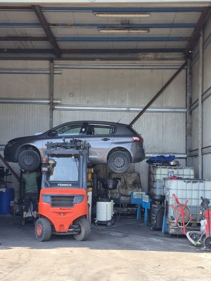 Un chariot élévateur soulève une voiture grise à l'intérieur d'un atelier. Divers équipements et conteneurs sont visibles.
