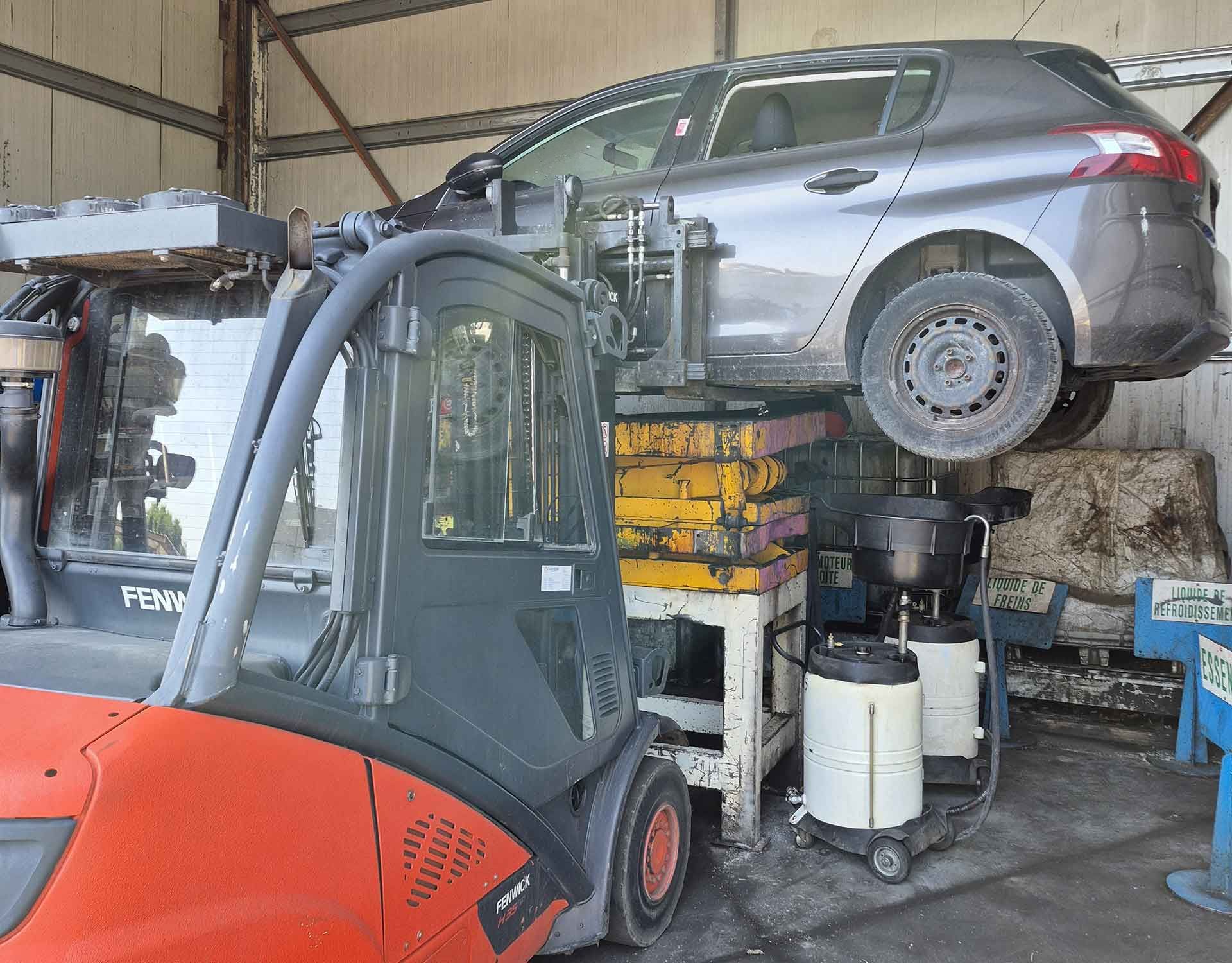 Un chariot élévateur orange soulève une voiture grise dans une casse.