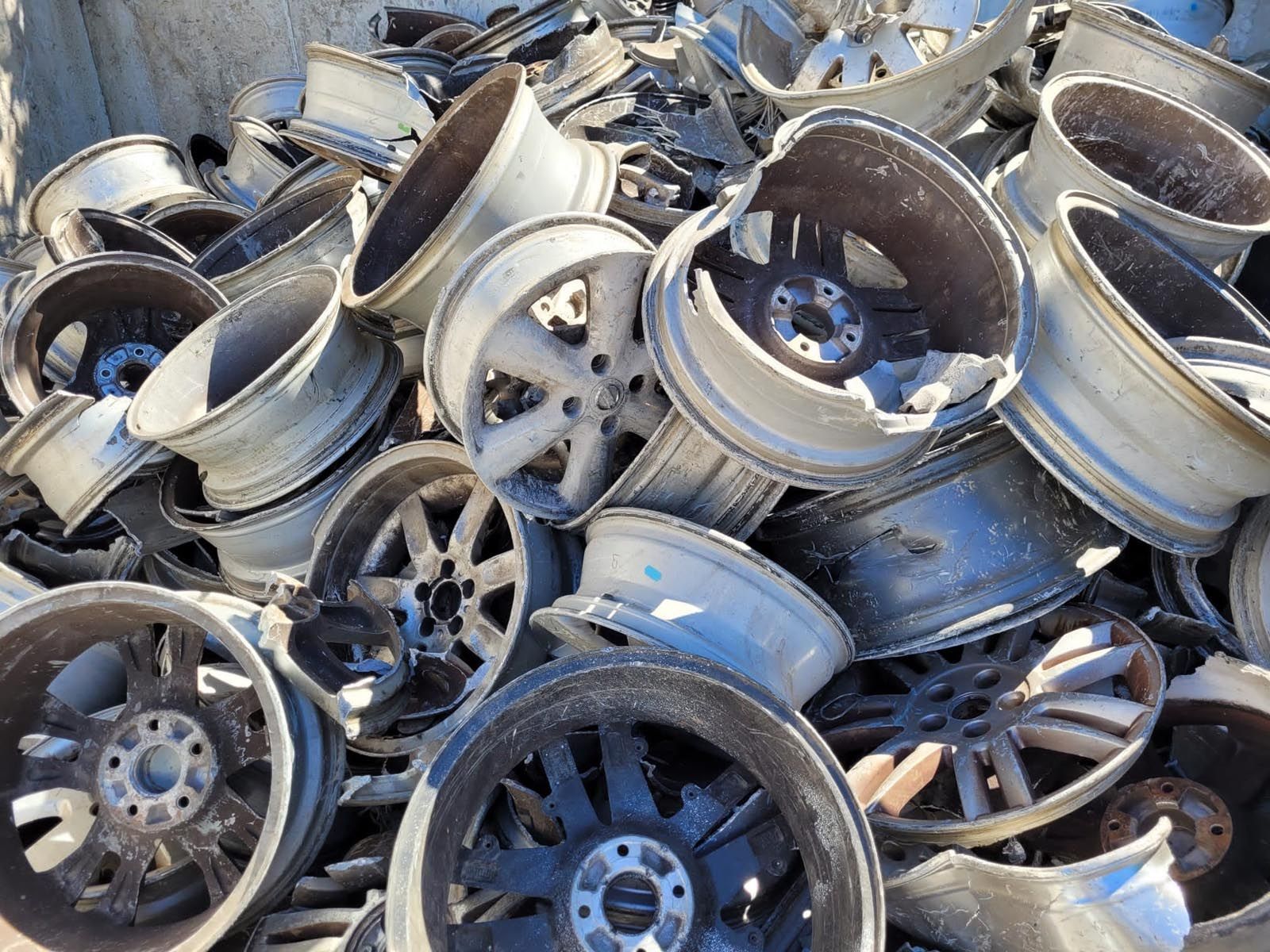 Un tas de jantes de voiture en alliage endommagées, principalement argentées et noires, dans une casse.