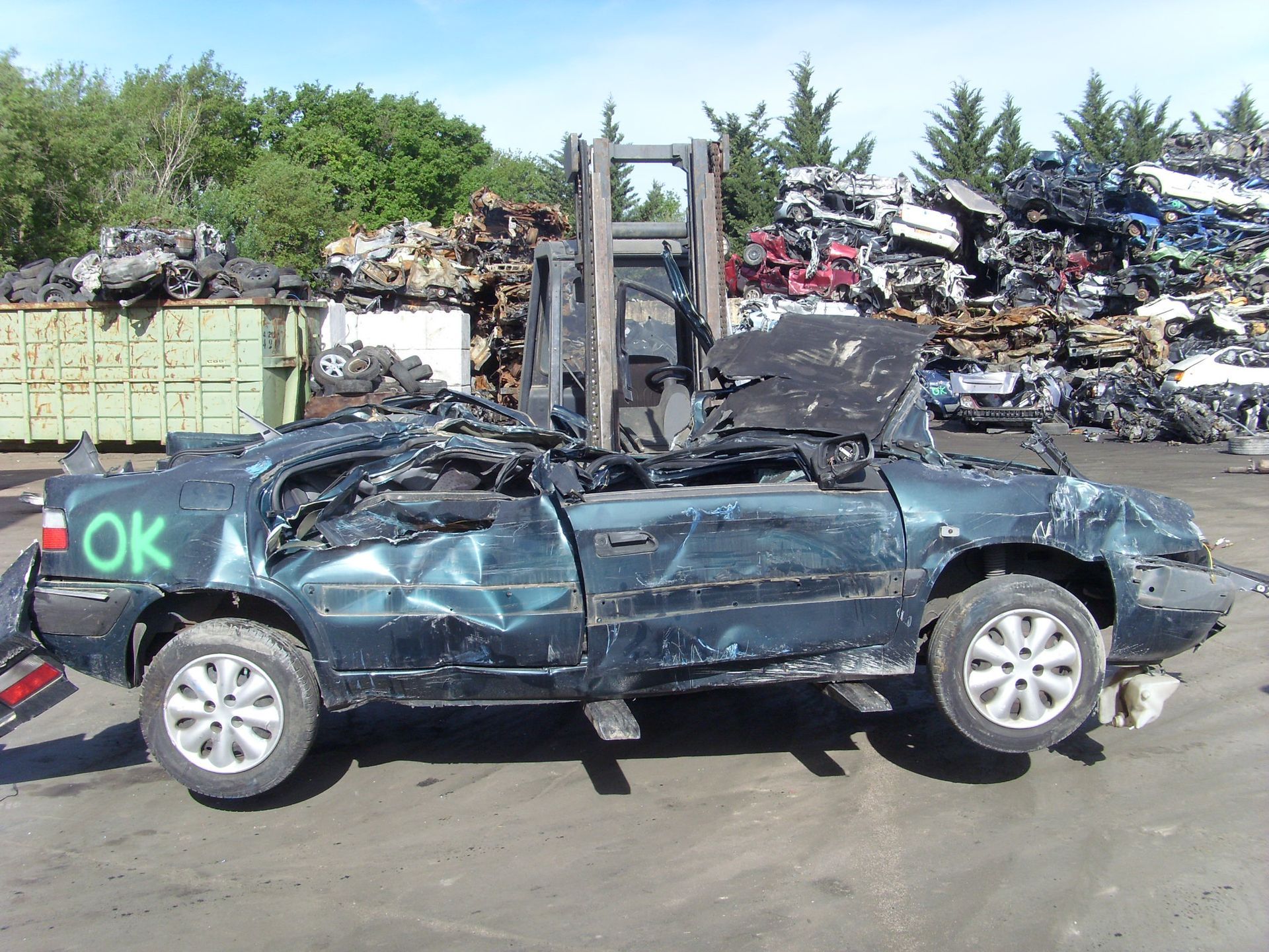 Une voiture turquoise écrasée, portant l'inscription OK, se trouve dans une casse et est soulevée par un chariot élévateur.