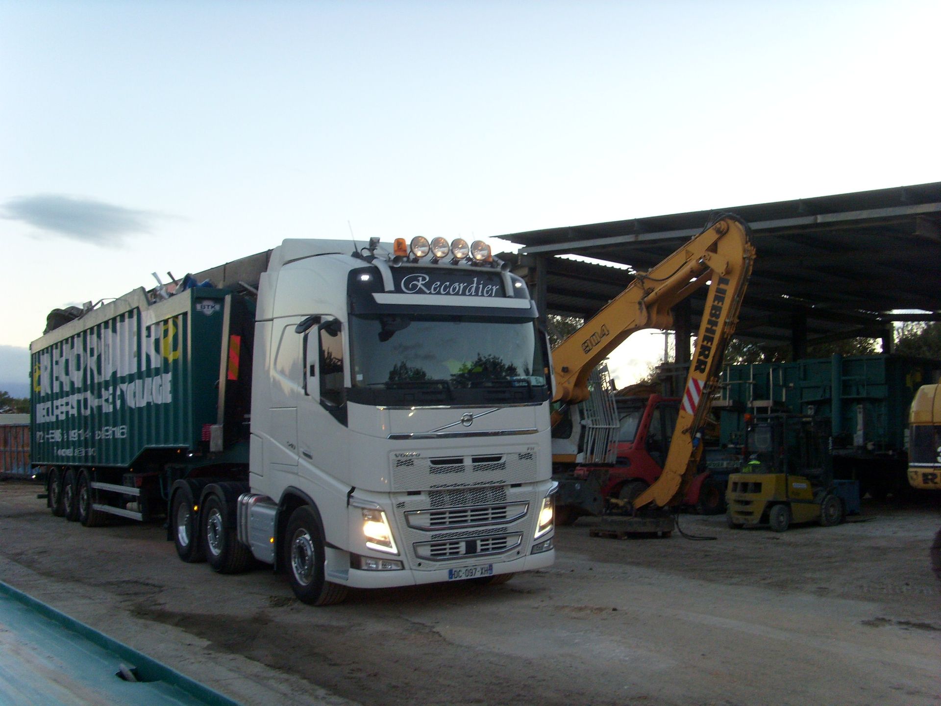 Un semi-remorque blanc transportant des débris est stationné près d'un entrepôt contenant du matériel de construction.