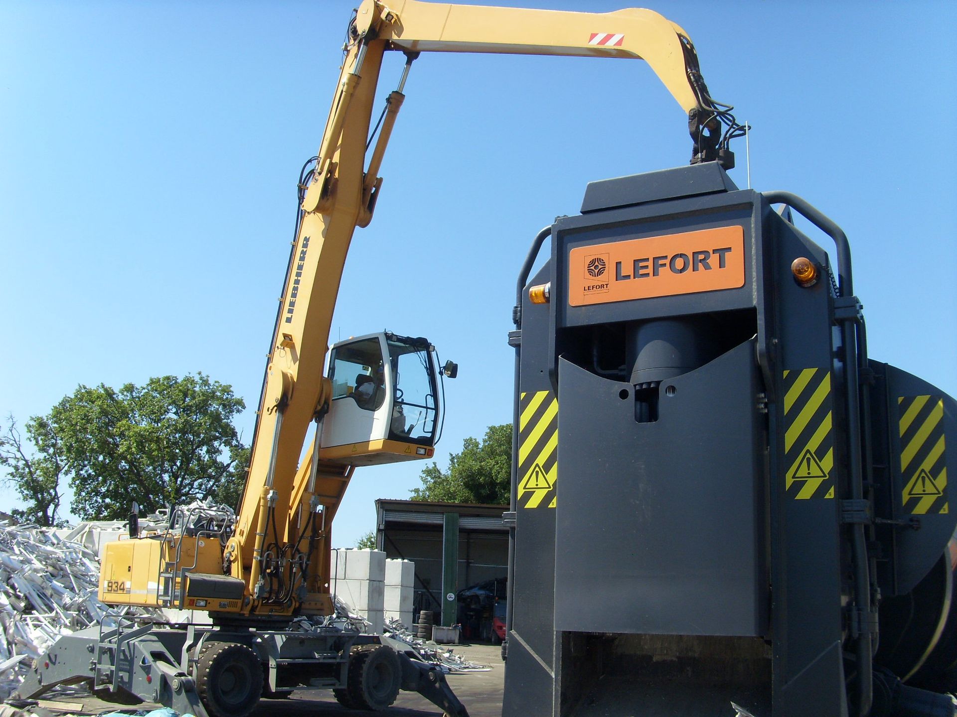 Une pelle mécanique jaune alimente une cisaille à ferraille Lefort noire. En extérieur, par une journée ensoleillée.