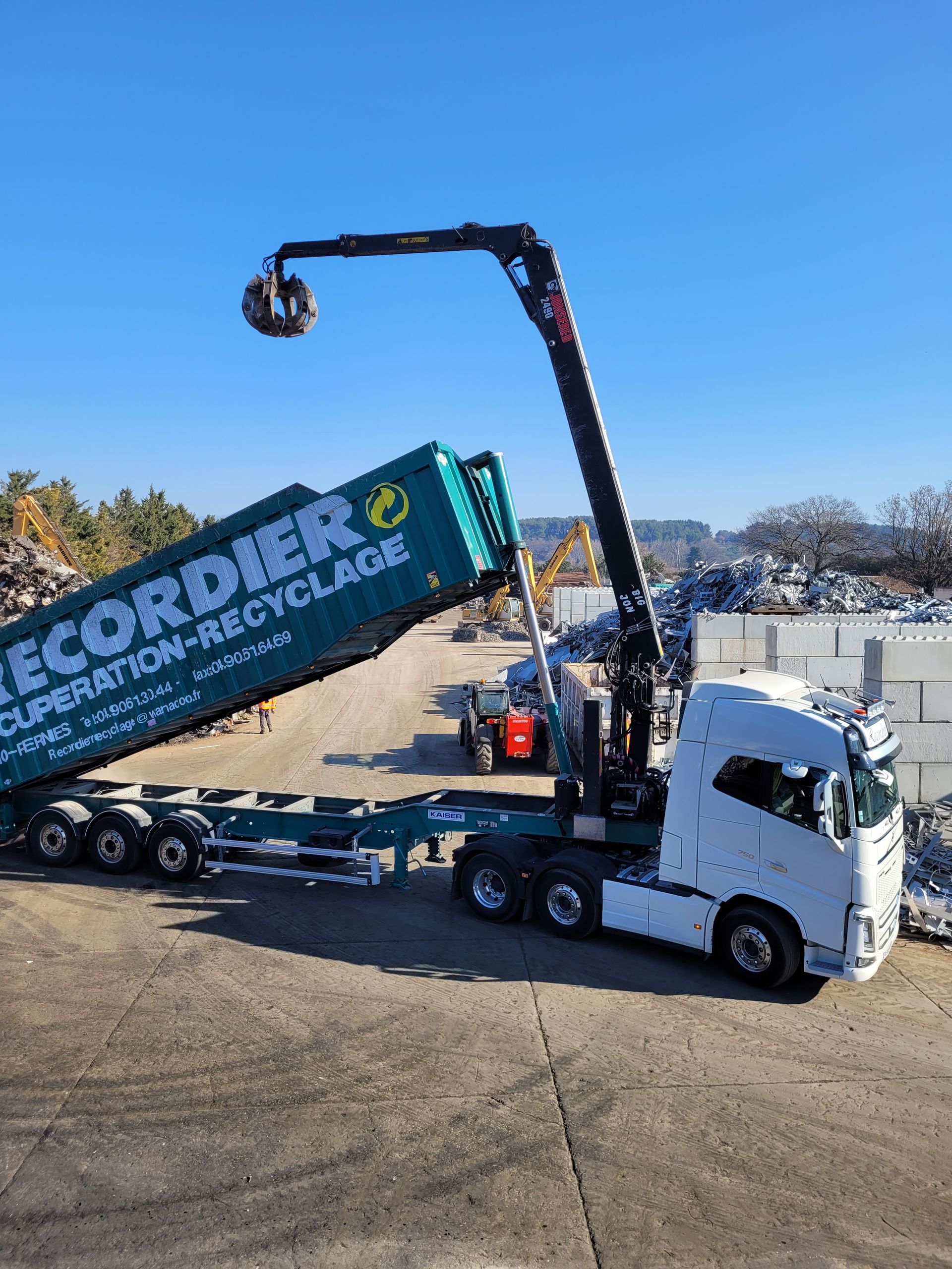 Un camion-grue RECORDIER RECYCLAGE soulève une boule de métal dans un centre de recyclage ; un conteneur est en cours de chargement.