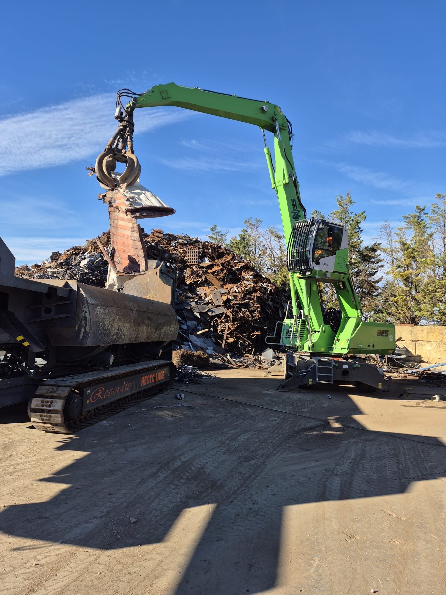 Un centre de recyclage de métaux avec des piles de ferraille, des blocs de béton et une grue sous un ciel bleu azur.