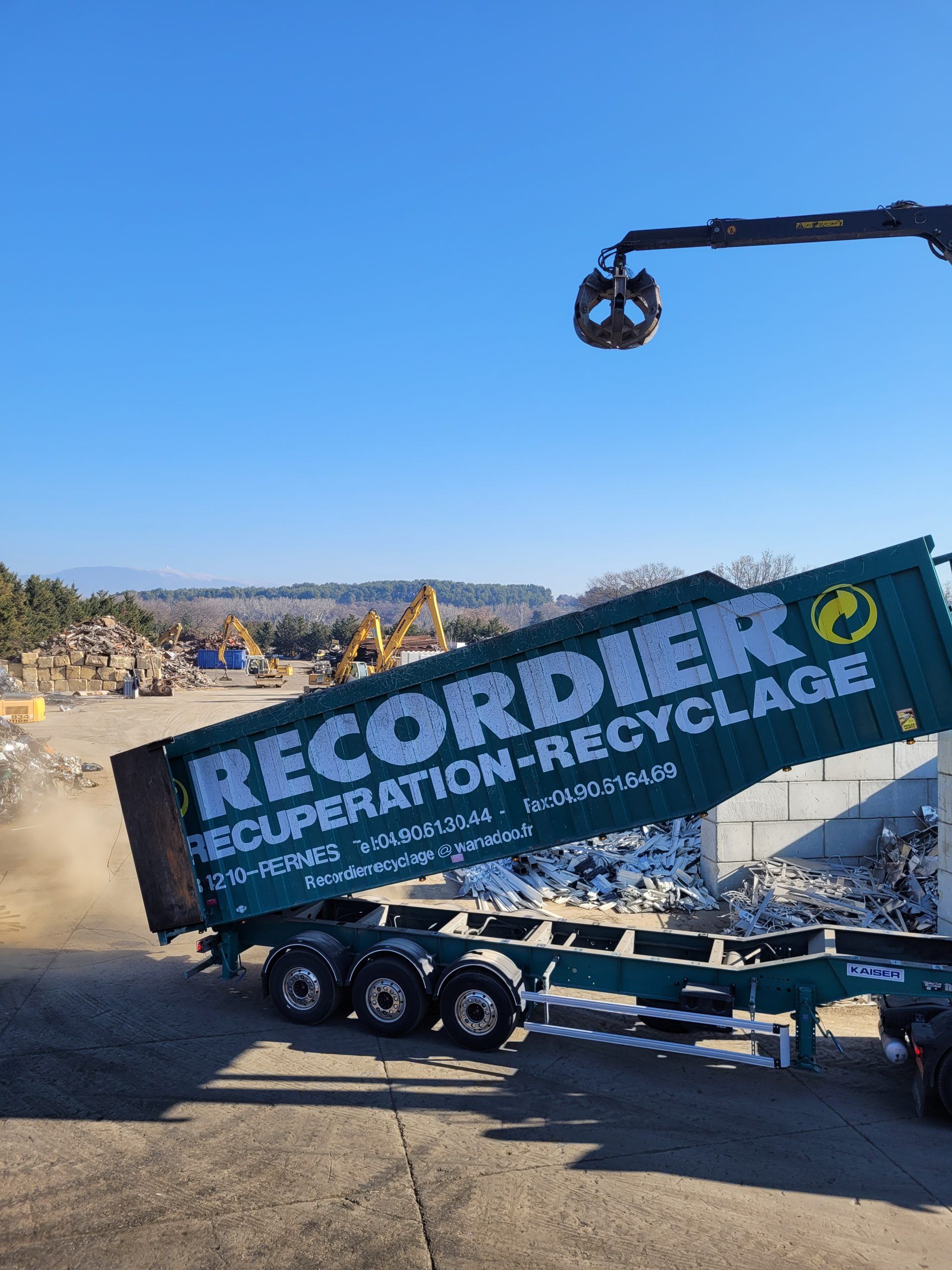 Un camion de recyclage vert décharge des débris dans une installation sous un ciel bleu, une grue au-dessus.