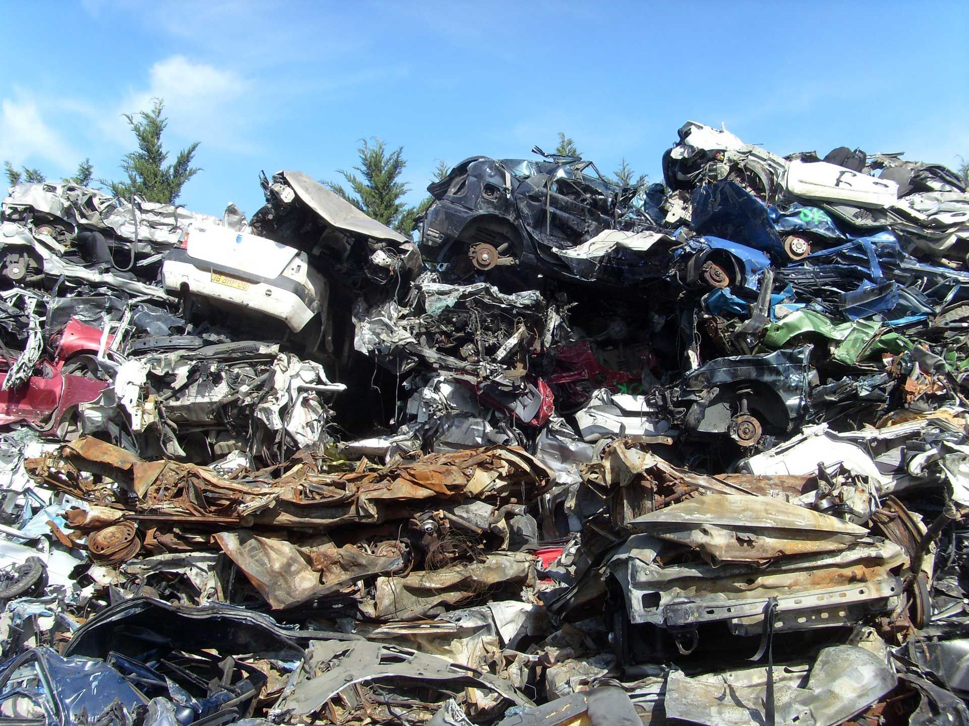 Un tas de voitures broyées dans une casse, sous un ciel bleu, des débris métalliques de couleurs variées.