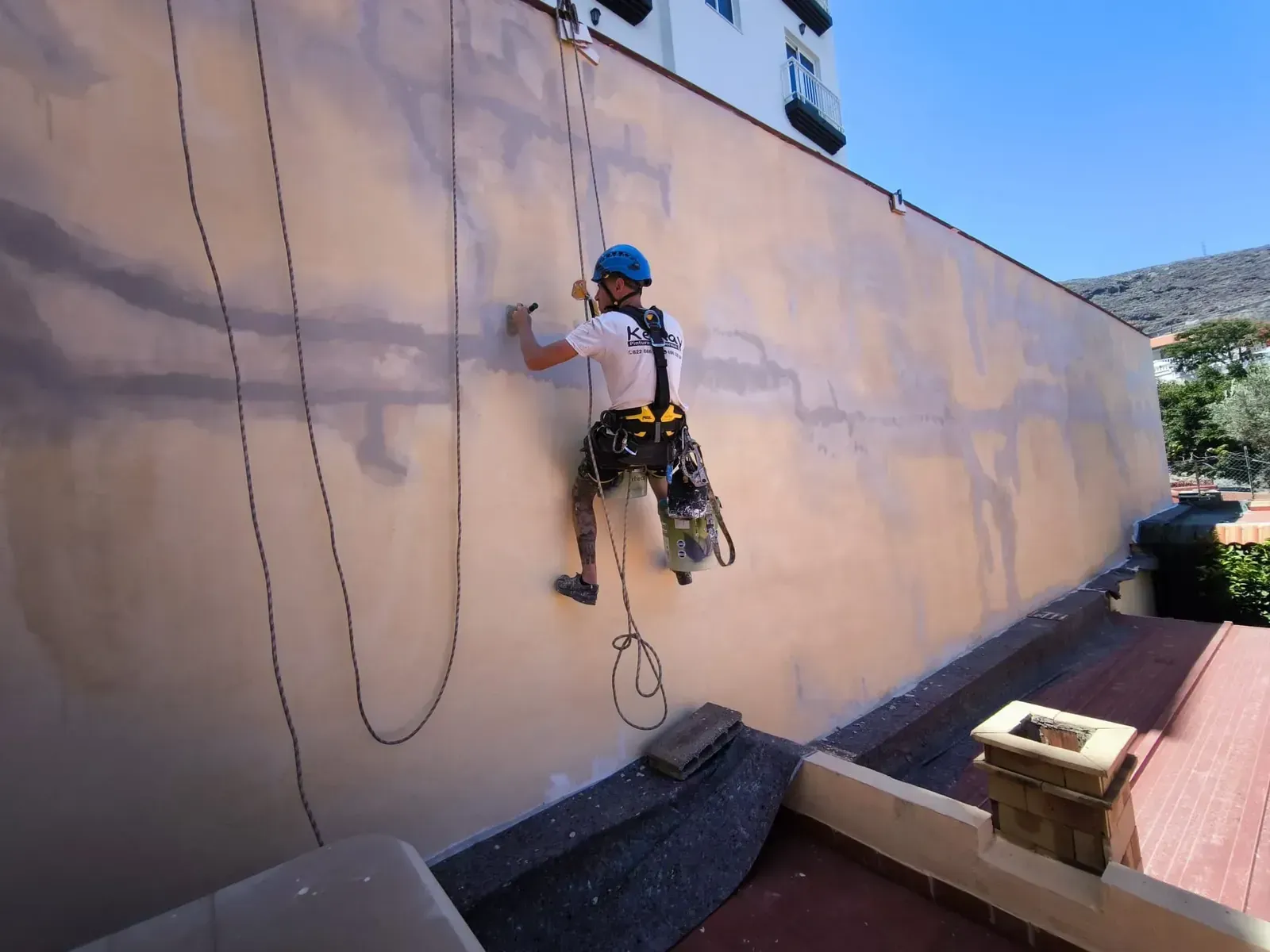 Un escalador desciende en rápel por una pared de color beige, con casco y arnés azules, y cuerdas colgando a su lado.
