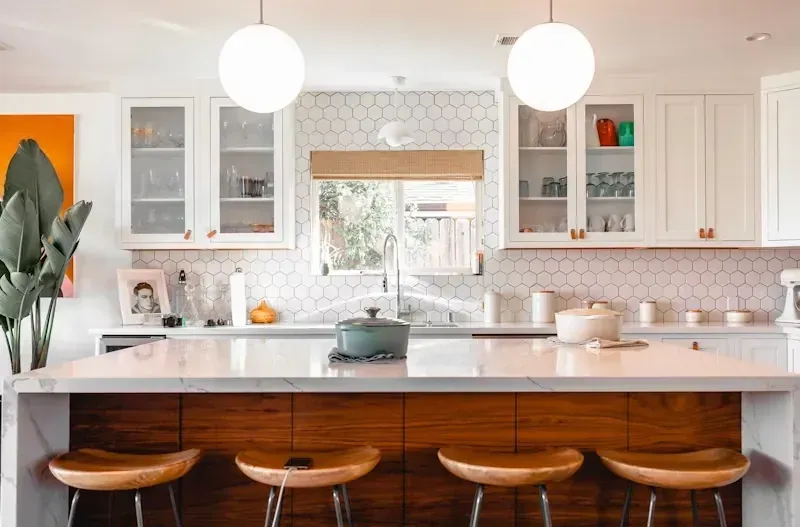 Cocina moderna y luminosa con armarios blancos, isla de madera, lámparas colgantes y tres taburetes.