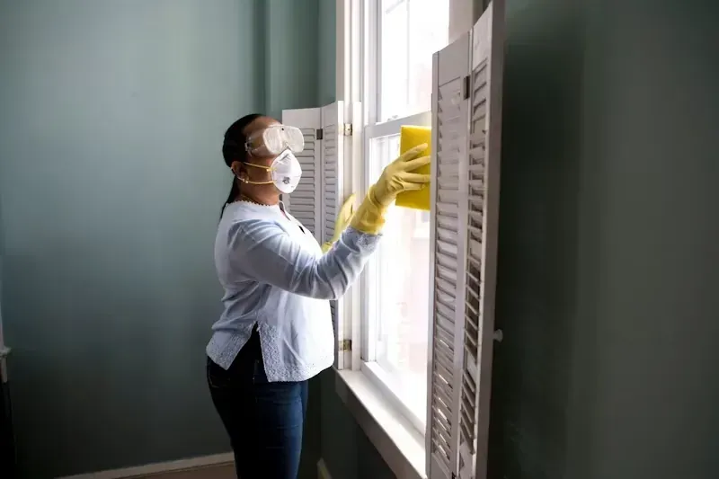 Una persona con mascarilla y guantes abre las persianas de una ventana en una habitación iluminada por el sol.