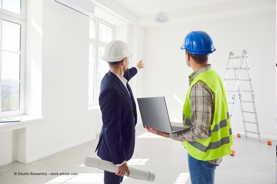 Ein Mann und ein Bauarbeiter schauen in einem leeren Raum auf einen Laptop.