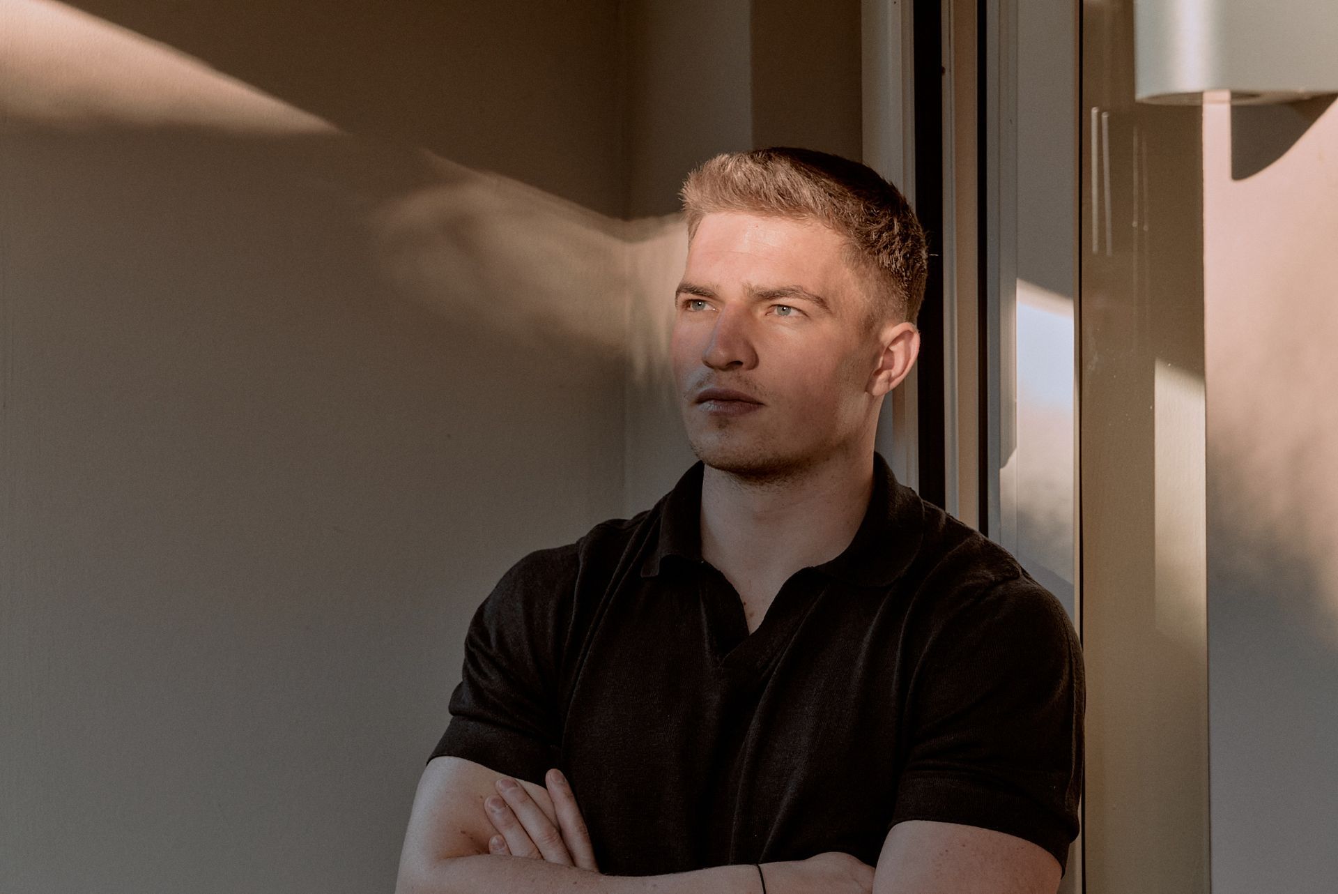 Eine Person mit kurzen Haaren und einem schwarzen Poloshirt steht mit verschränkten Armen.
