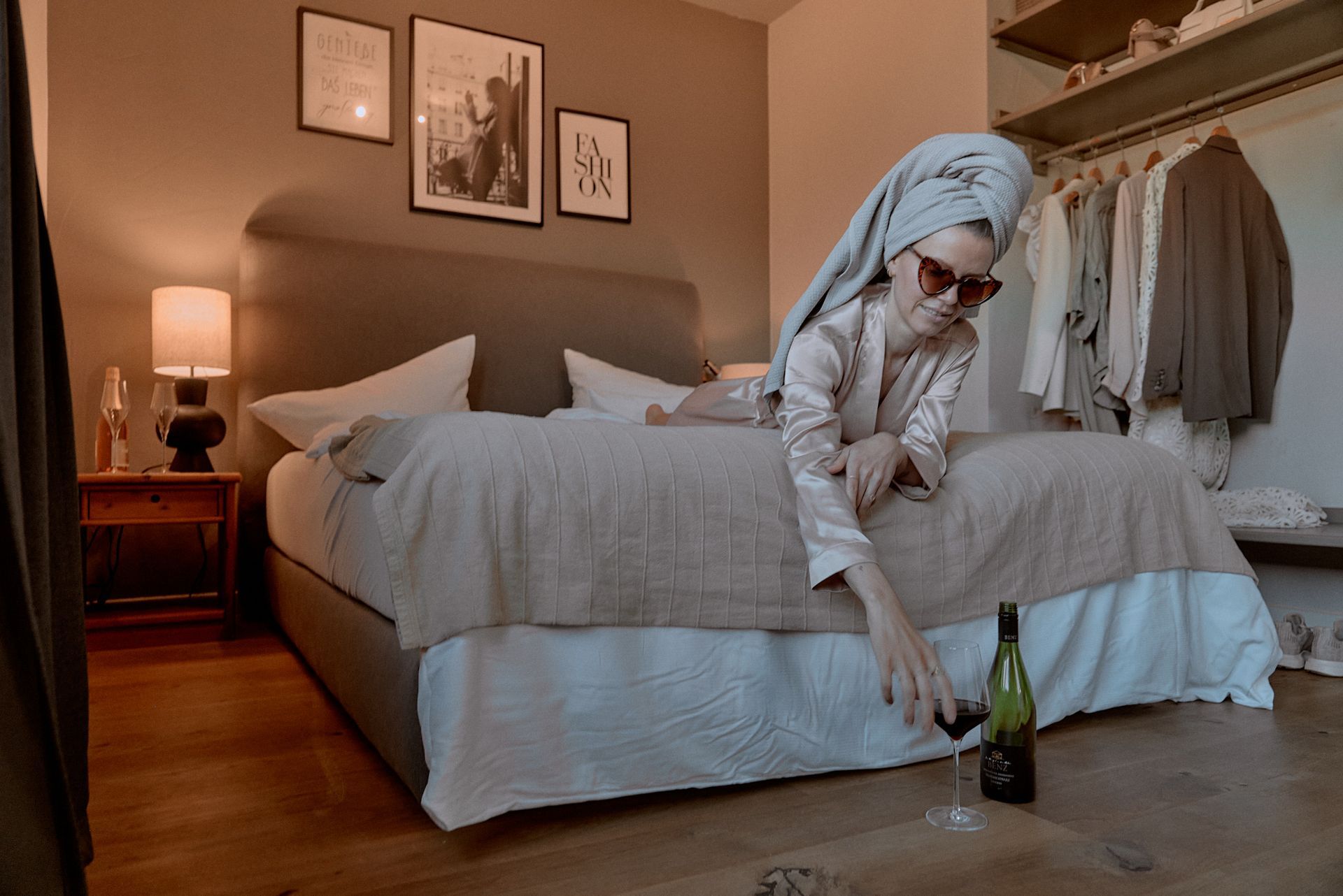 Eine Frau liegt mit einem Handtuch um den Kopf und einer Flasche Wein auf einem Bett.