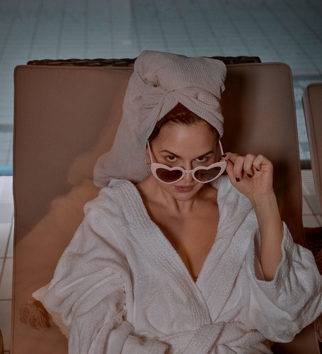 Eine Frau in Bademantel und Handtuch, die eine herzförmige Sonnenbrille trägt, liegt am Pool.