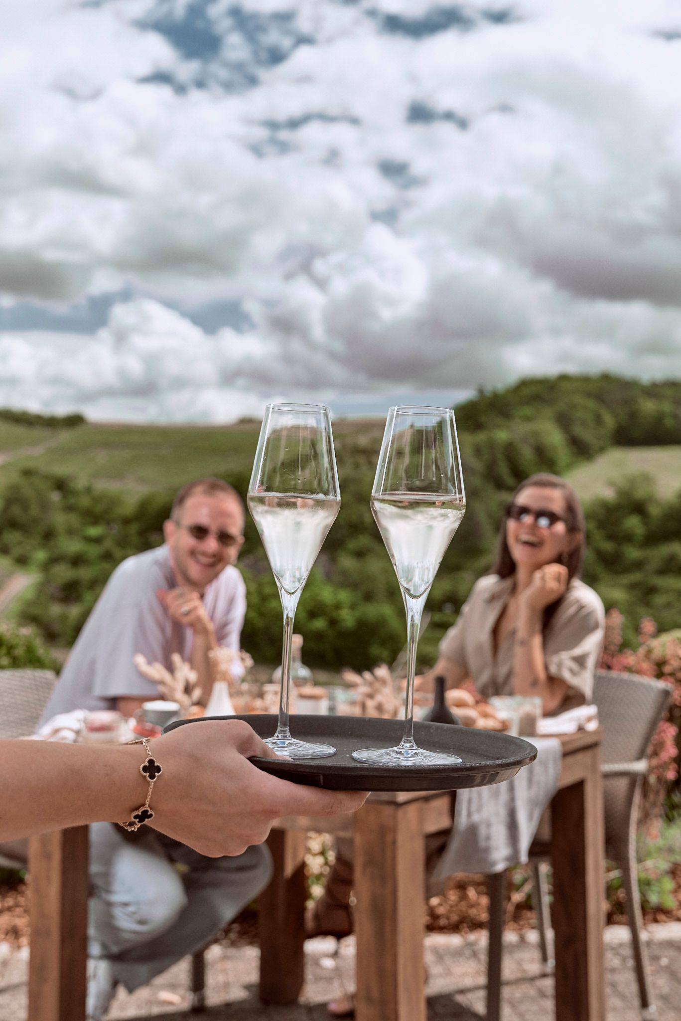 Person mit Champagnertablett, zwei Personen sitzen draußen an einem Tisch, im Hintergrund Weinberg.