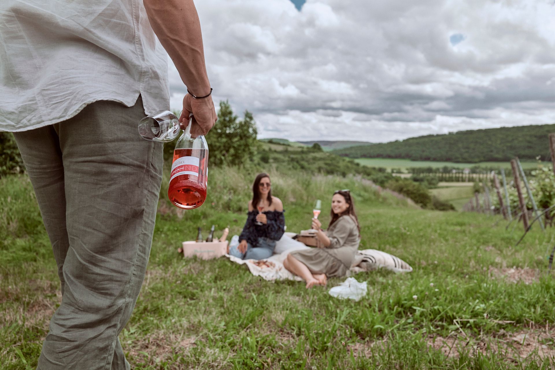 Ein Mann hält eine Flasche Wein, während zwei Frauen im Gras ein Picknick machen.