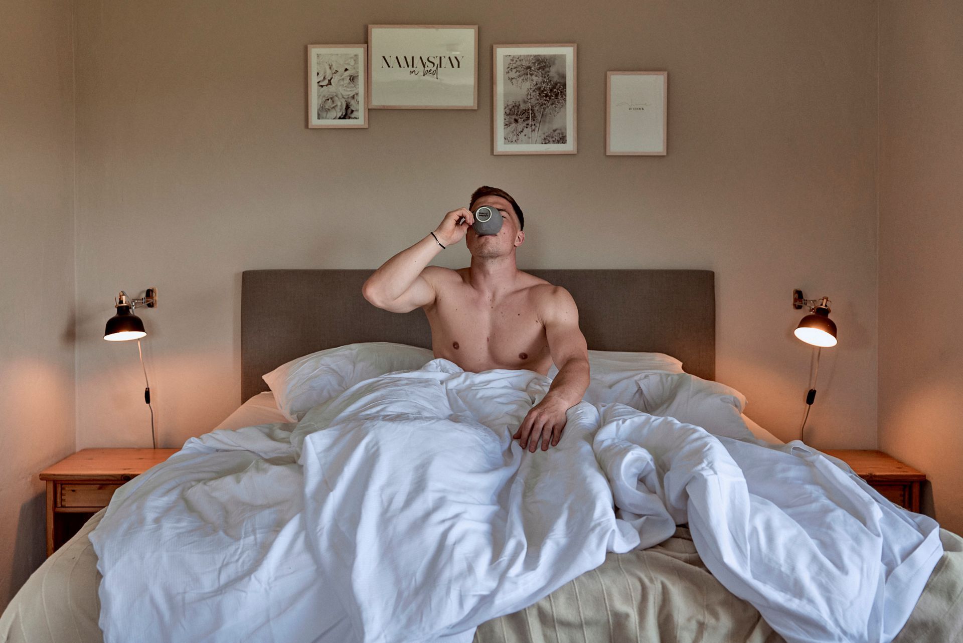 Ein Mann ohne Hemd sitzt auf einem Bett und trinkt aus einer Flasche.