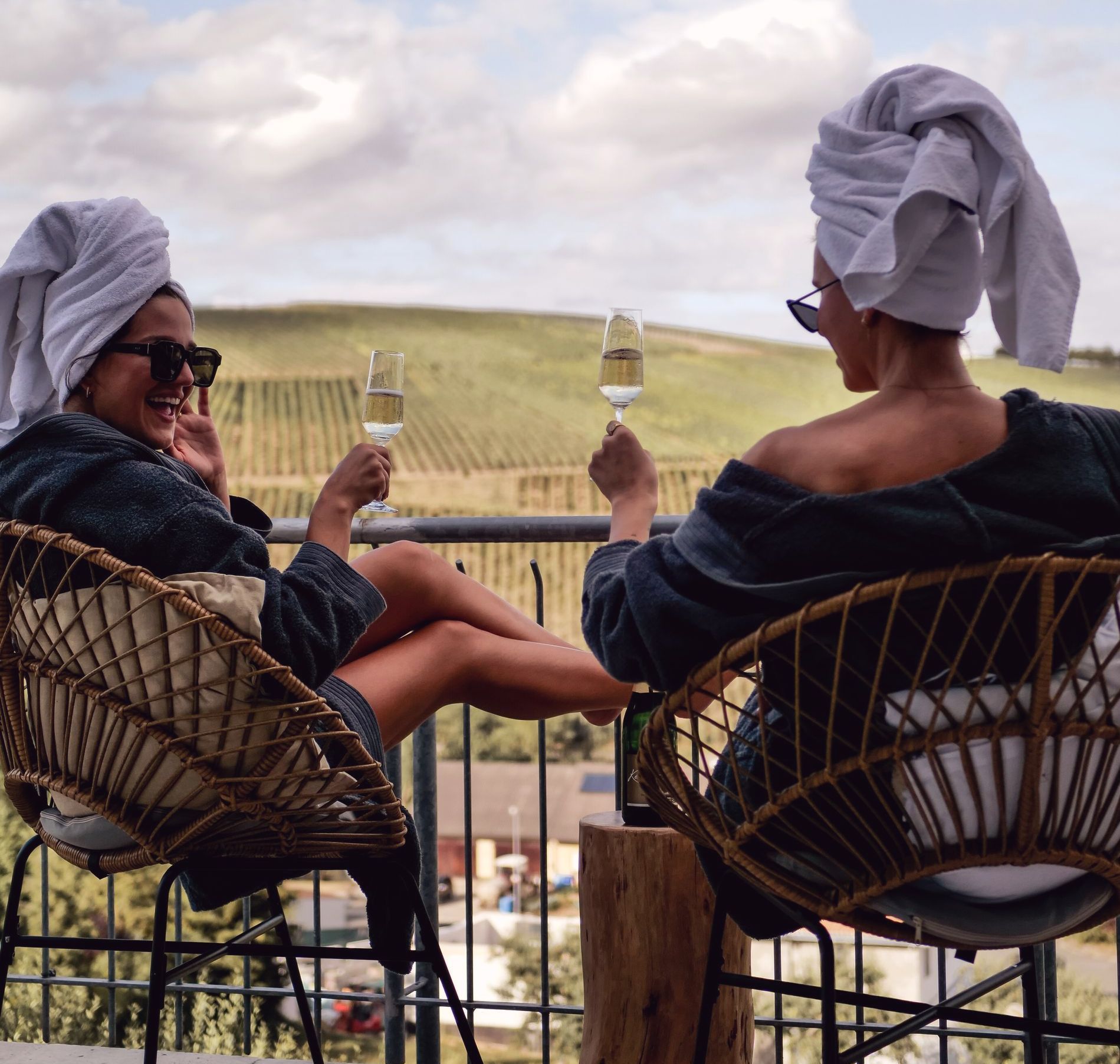 Zwei Personen in Roben und Kopftüchern stoßen auf einem Balkon mit Blick auf die Weinberge mit Champagner an.