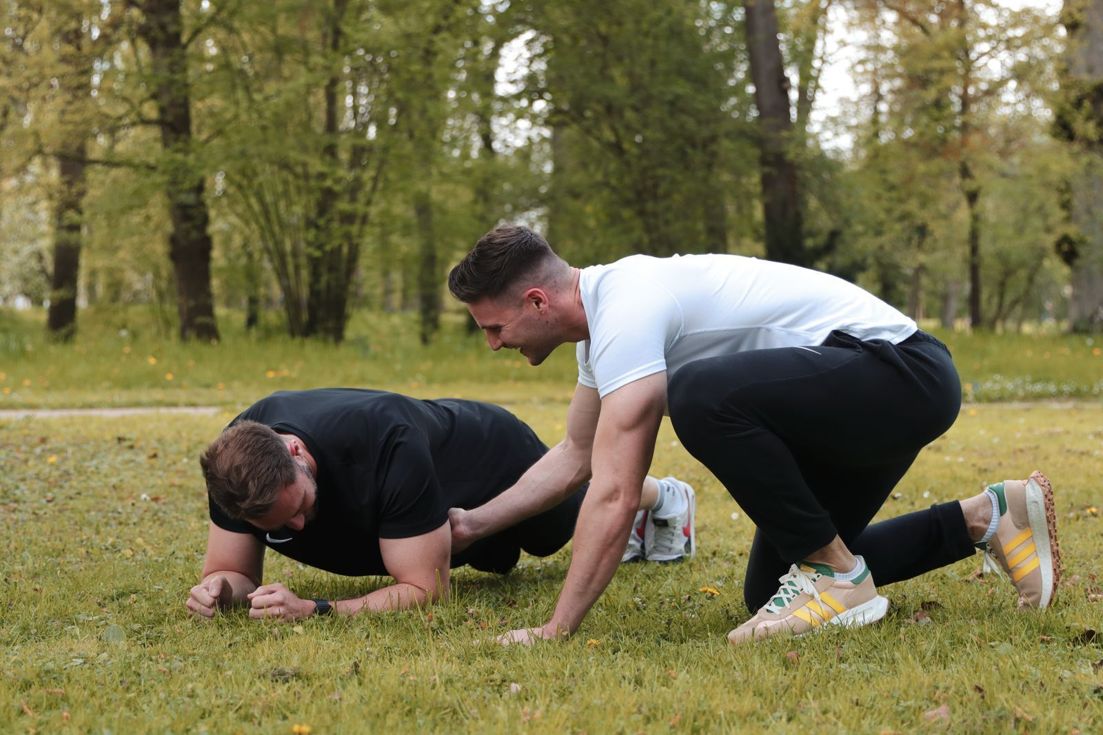 Zwei Männer trainieren im Freien: einer in der Plank-Position, der andere hilft ihm; grasbewachsene Parklandschaft.