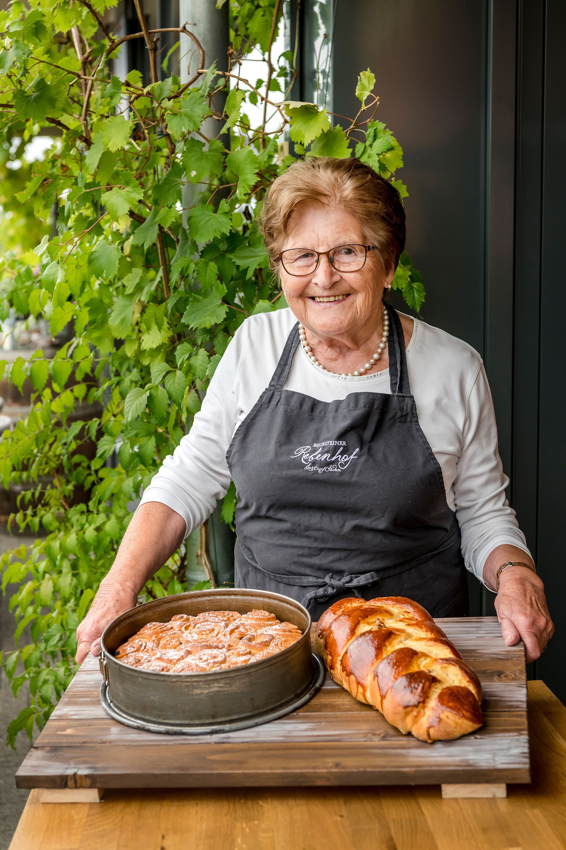 Eine ältere Frau steht neben einem Kuchen und einem Laib Brot auf einem Holztisch.