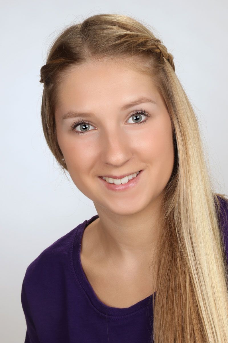Blonde Person lächelt, trägt ein lila Hemd und hat einen Zopf über der Stirn.