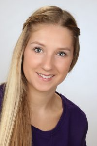 Blonde Person lächelt, trägt ein lila Hemd und hat einen Zopf über der Stirn.