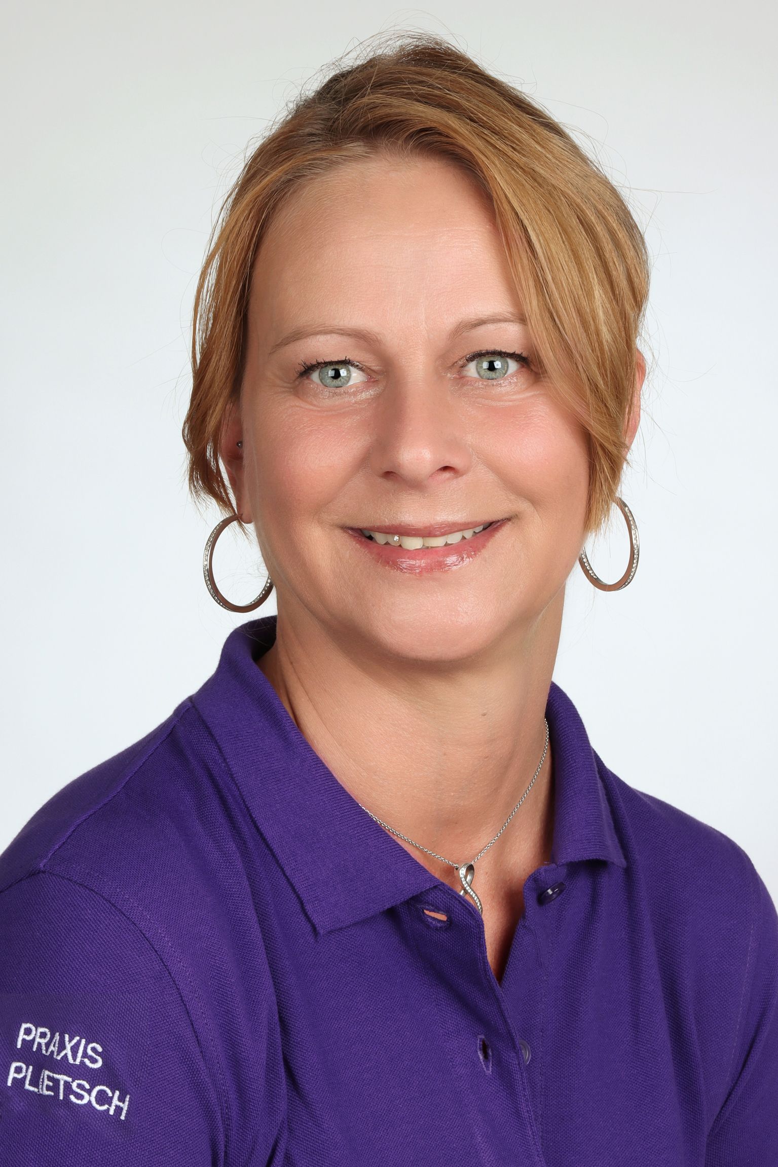 Eine Frau mit blonden Haaren, die ein lila Poloshirt trägt, lächelt vor einem neutralen Hintergrund.