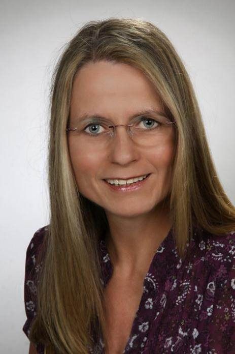 Frau mit langen blonden Haaren, Brille und lila geblümtem Hemd, lächelt.