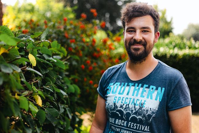 Ein Mann mit Bart und blauem T-Shirt steht vor einem Busch.