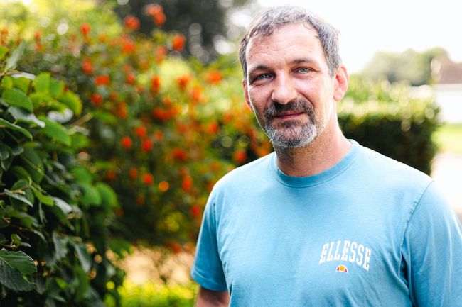 Ein Mann mit Bart und blauem T-Shirt steht vor einem Busch.