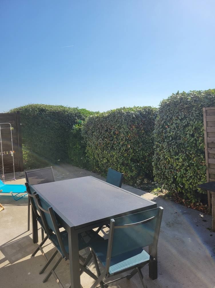 Patio avec une table noire, des chaises et de hautes haies vertes sous un ciel bleu.