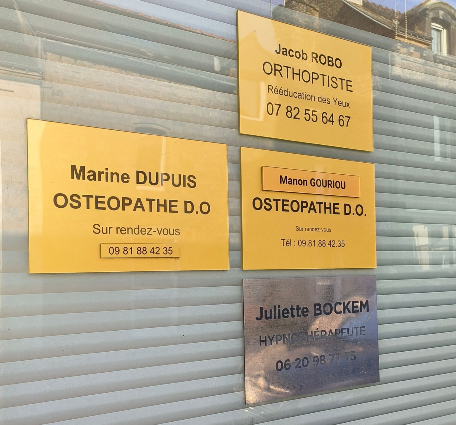 Des panneaux jaunes sur une fenêtre : Jacob Robo (orthoptiste), Marine Dupuis (ostéopathe), Marion GOUJON (ostéopathe) et Juliette Bockem (hypnothérapeute), avec leurs coordonnées.