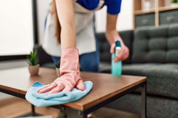 Person reinigt einen Holztisch mit einem blauen Tuch und einer Sprühflasche in einem Wohnzimmer.