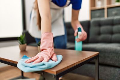 Person reinigt einen Holztisch mit einem blauen Tuch und einer Sprühflasche in einem Wohnzimmer.