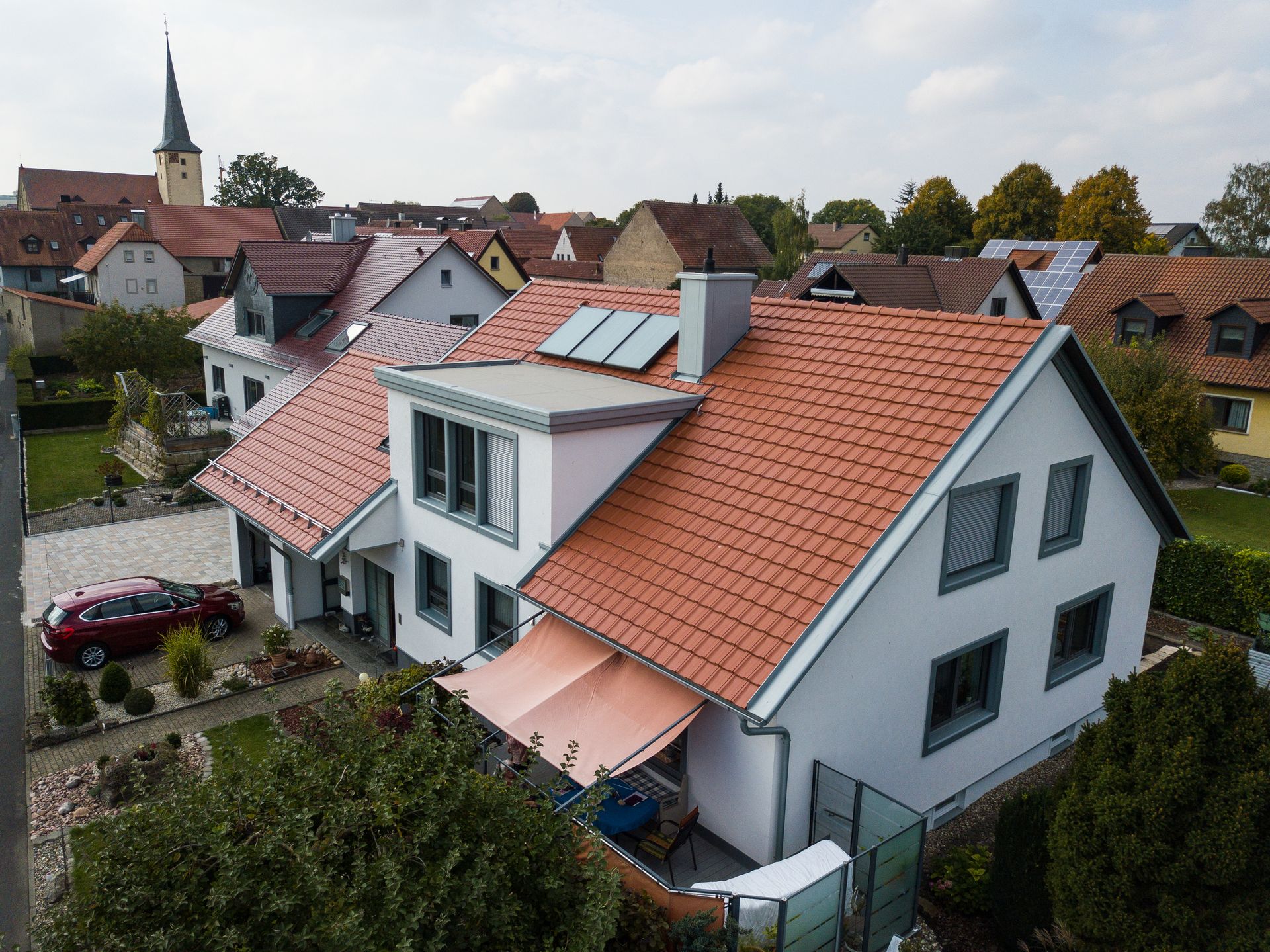 Luftaufnahme von weißen Häusern mit roten Ziegeldächern in einem Wohngebiet, im Hintergrund ein Kirchturm.
