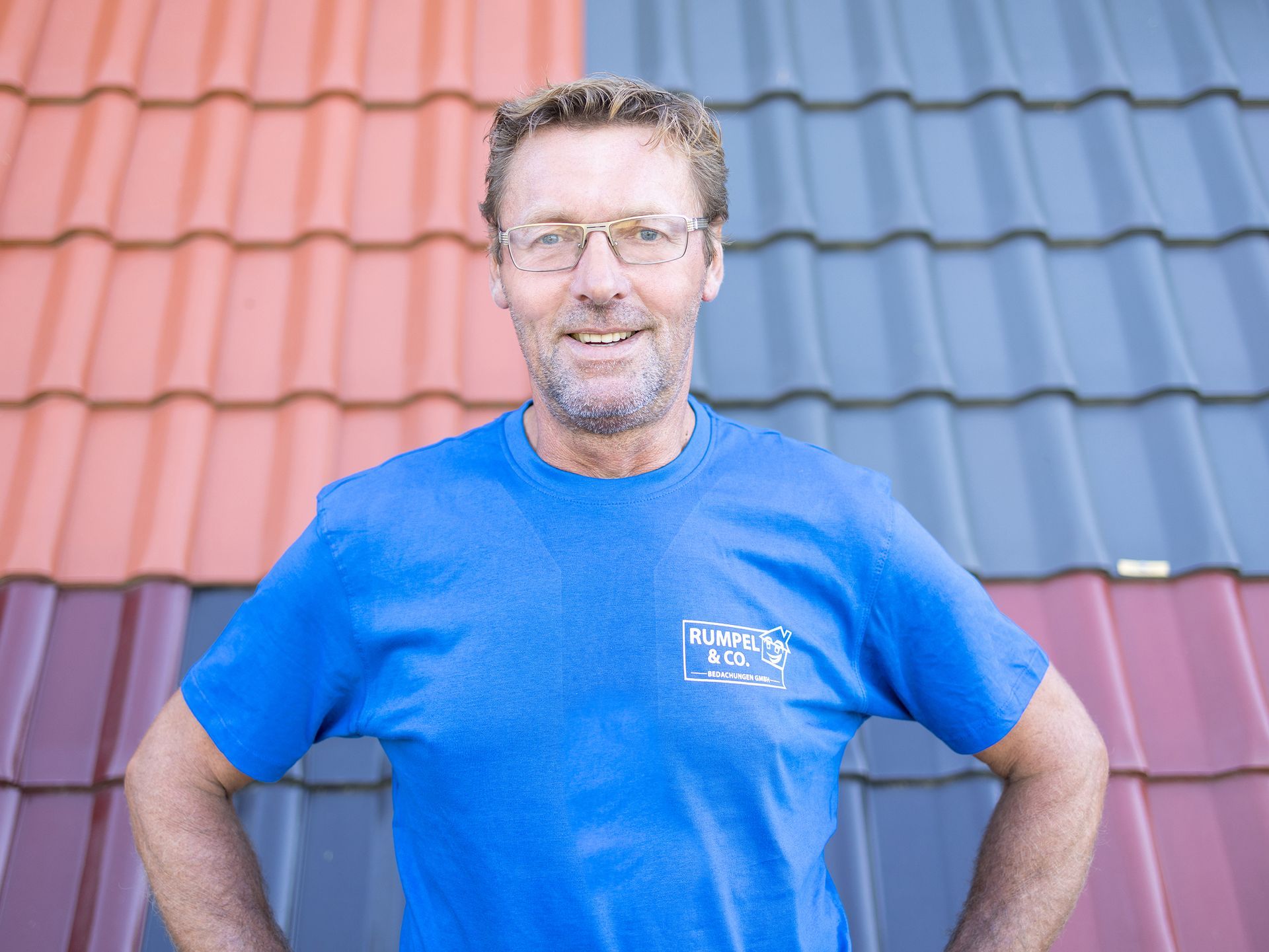 Ein Mann in blauem T-Shirt steht mit in die Hüften gestemmten Händen vor einer rot-grau gefliesten Wand.