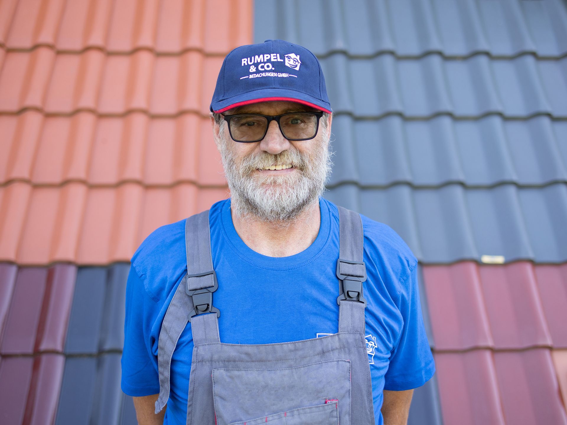 Ein lächelnder Mann in blauem Hemd steht vor einem rot-blauen Ziegeldach.