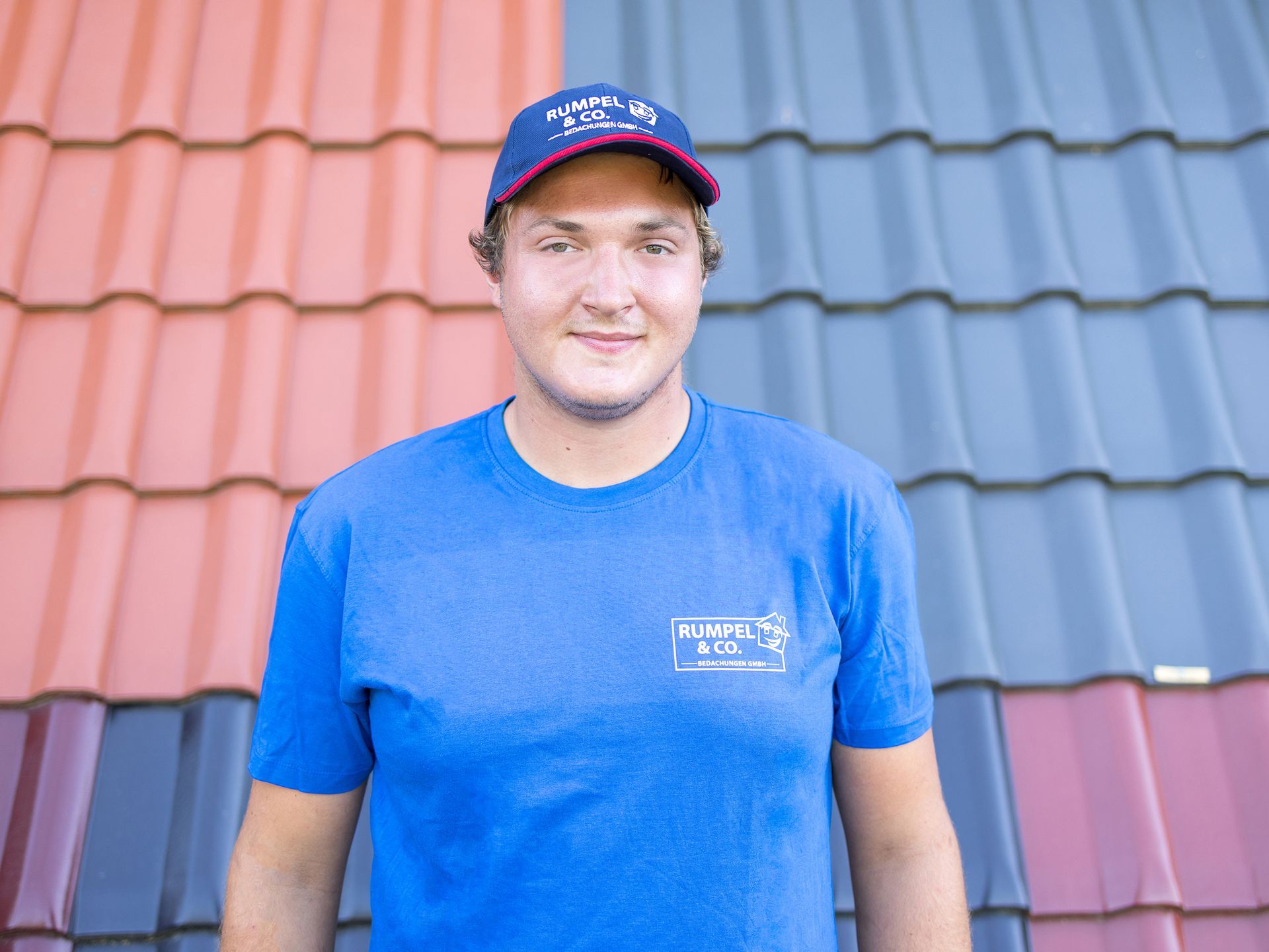 Ein Mann in einem blauen T-Shirt steht mit den Händen in den Hüften vor einem rot-grauen Ziegeldach.