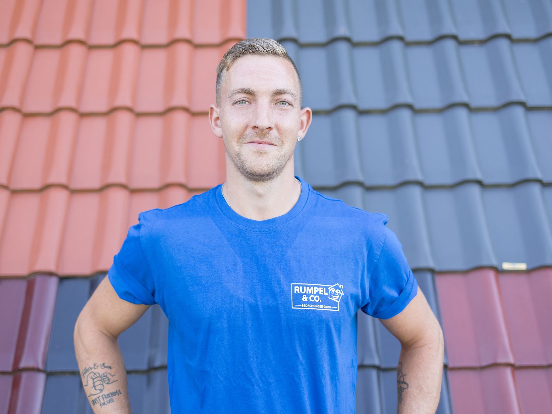 Ein Mann in blauem Hemd und Latzhose steht vor einem roten Ziegeldach; er trägt eine Kappe und eine Brille.