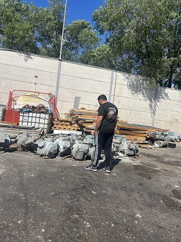 Una persona se encuentra de pie en un terreno baldío junto a montones de escombros y bolsas de basura cerca de un muro de hormigón y árboles.