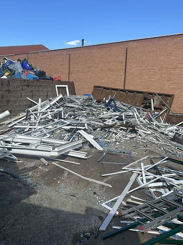 Montón de escombros metálicos blancos en un estacionamiento junto a una pared de ladrillos, con contenedores de basura al fondo.