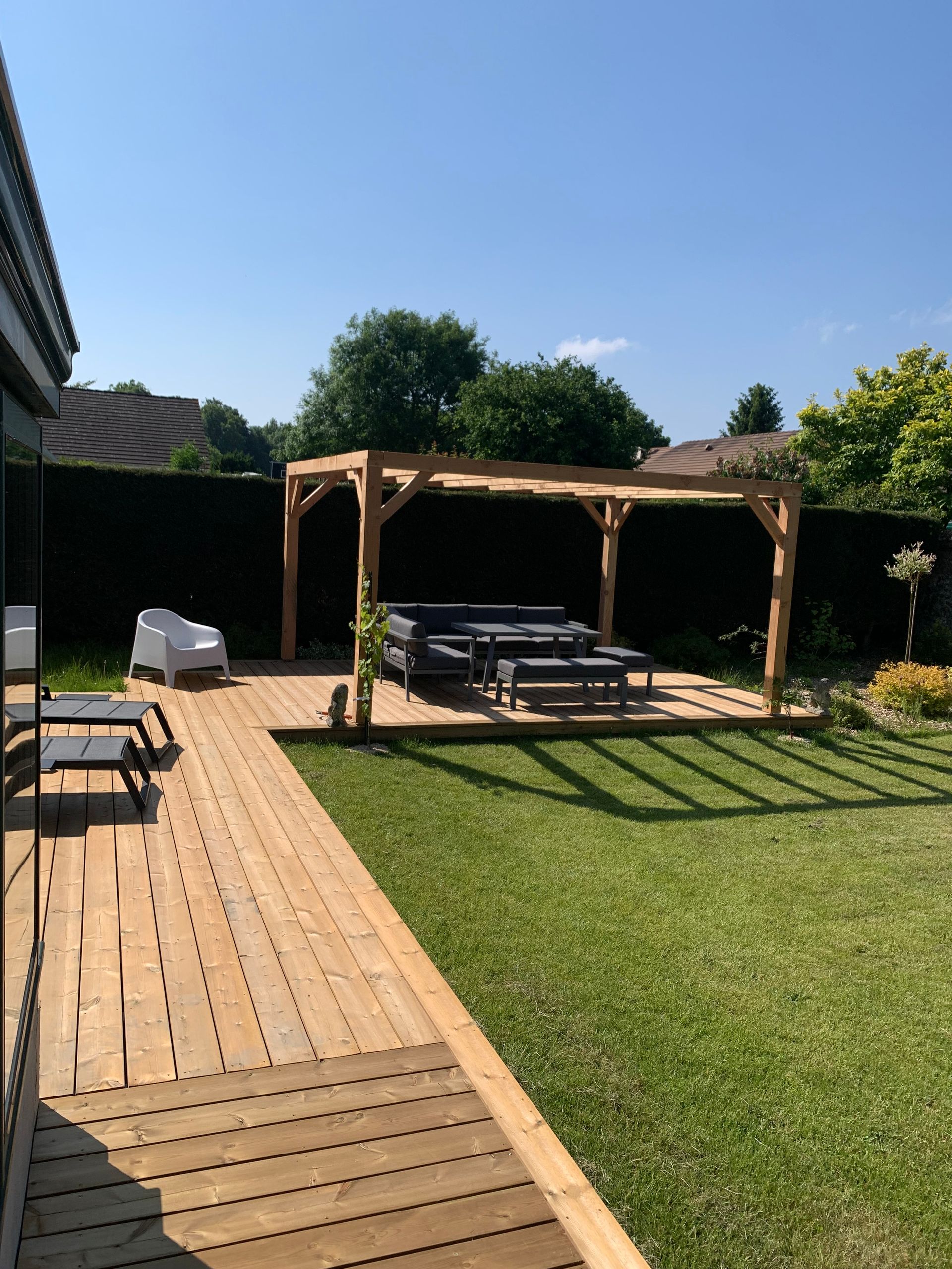 Terrasse en bois et pergola dans un jardin ensoleillé avec mobilier de jardin et pelouse verte.