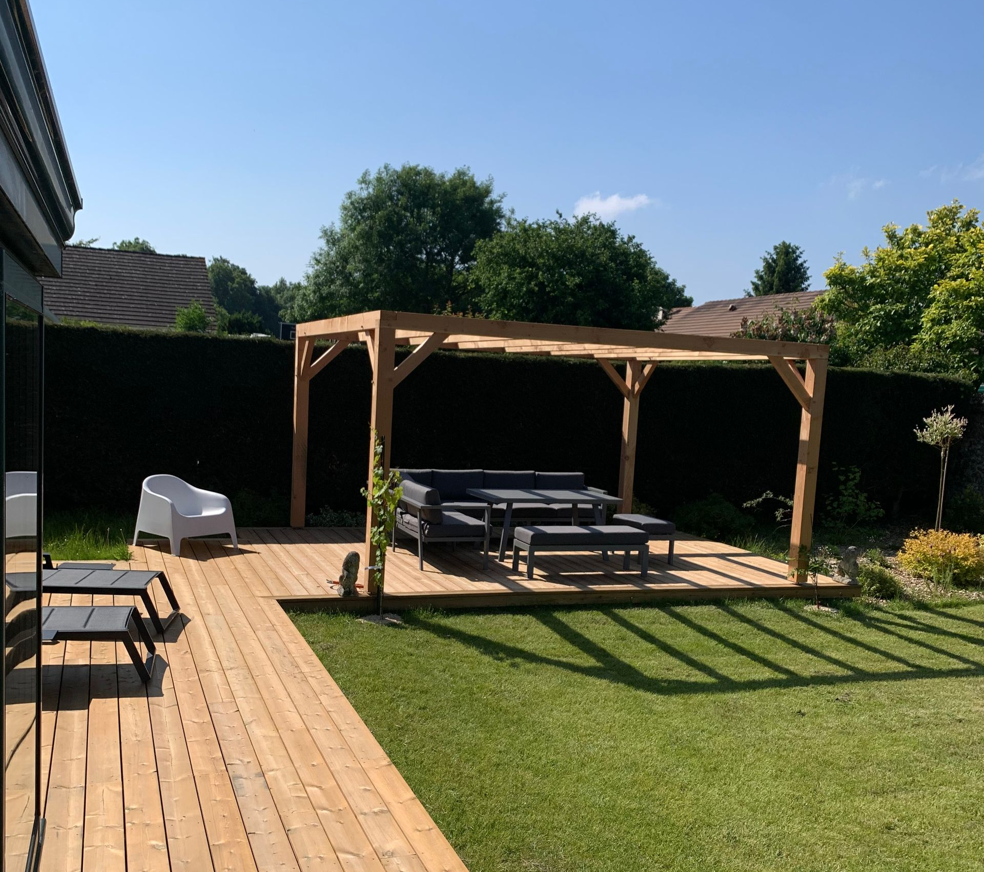 Pergola en bois avec sièges extérieurs sur une terrasse dans une cour arrière avec pelouse et haute haie.