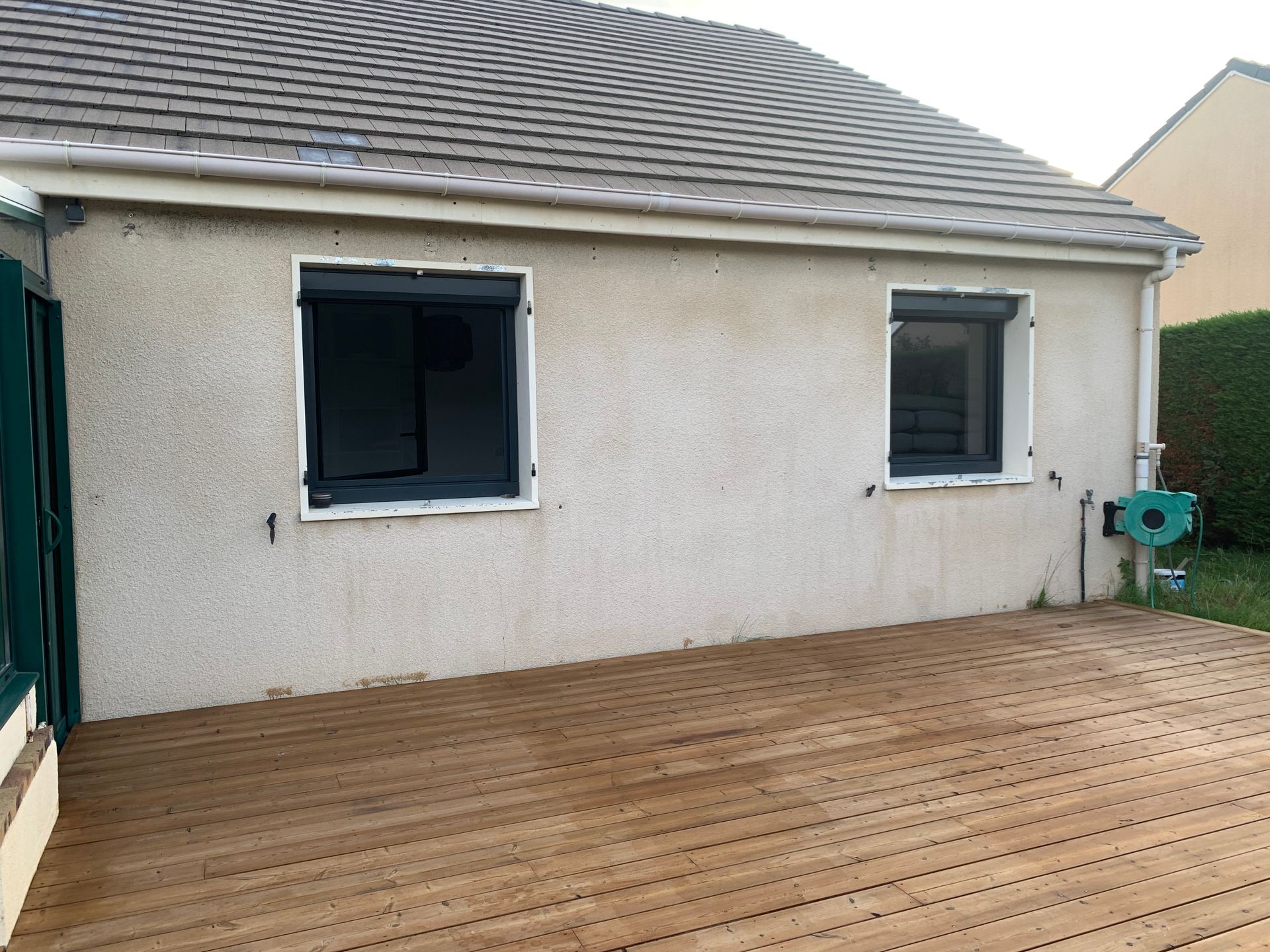 Vue extérieure d'une maison avec une terrasse en bois et deux fenêtres à volets roulants.