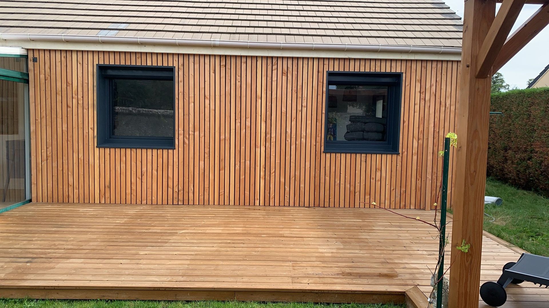 Maison en bois avec deux fenêtres à cadre foncé sur une terrasse en bois.
