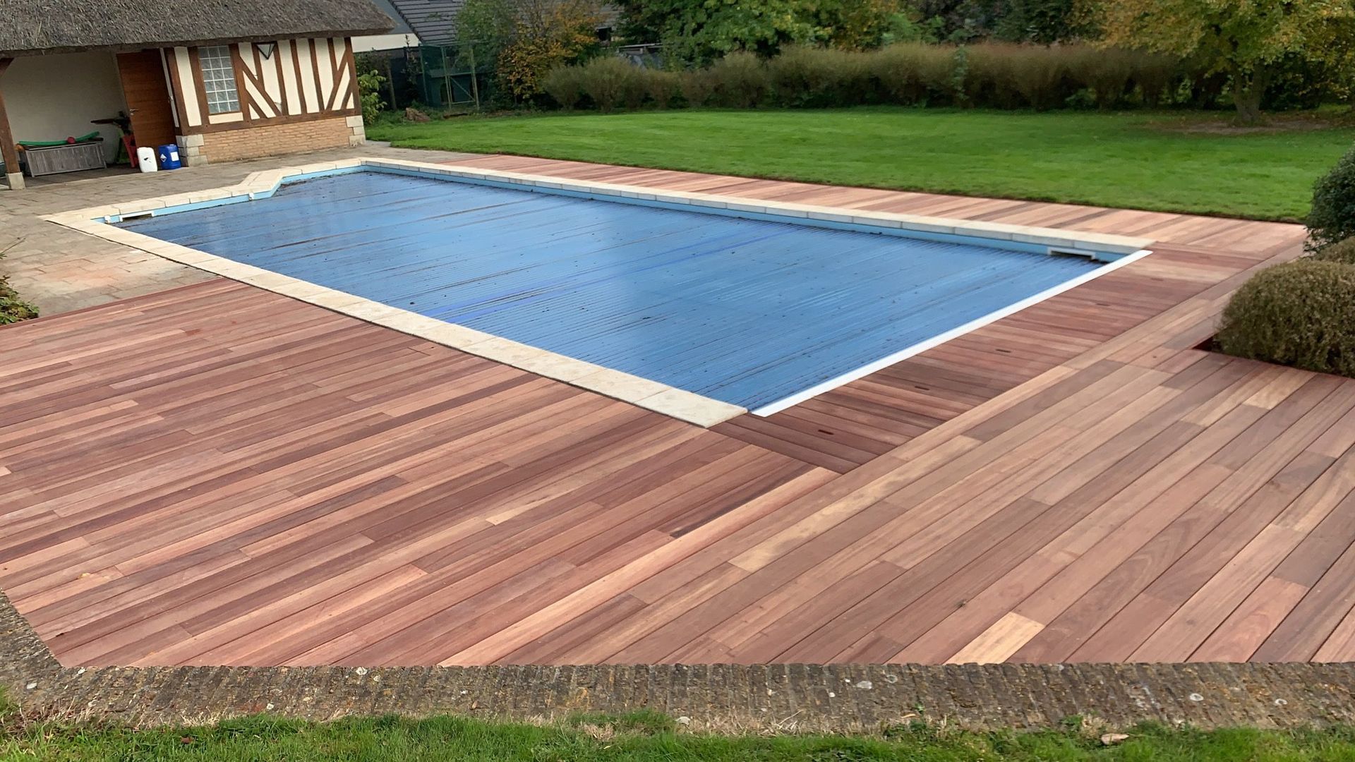 Piscine rectangulaire avec protection bleue, entourée d'une terrasse en bois et d'une pelouse, à côté d'un bâtiment en briques.