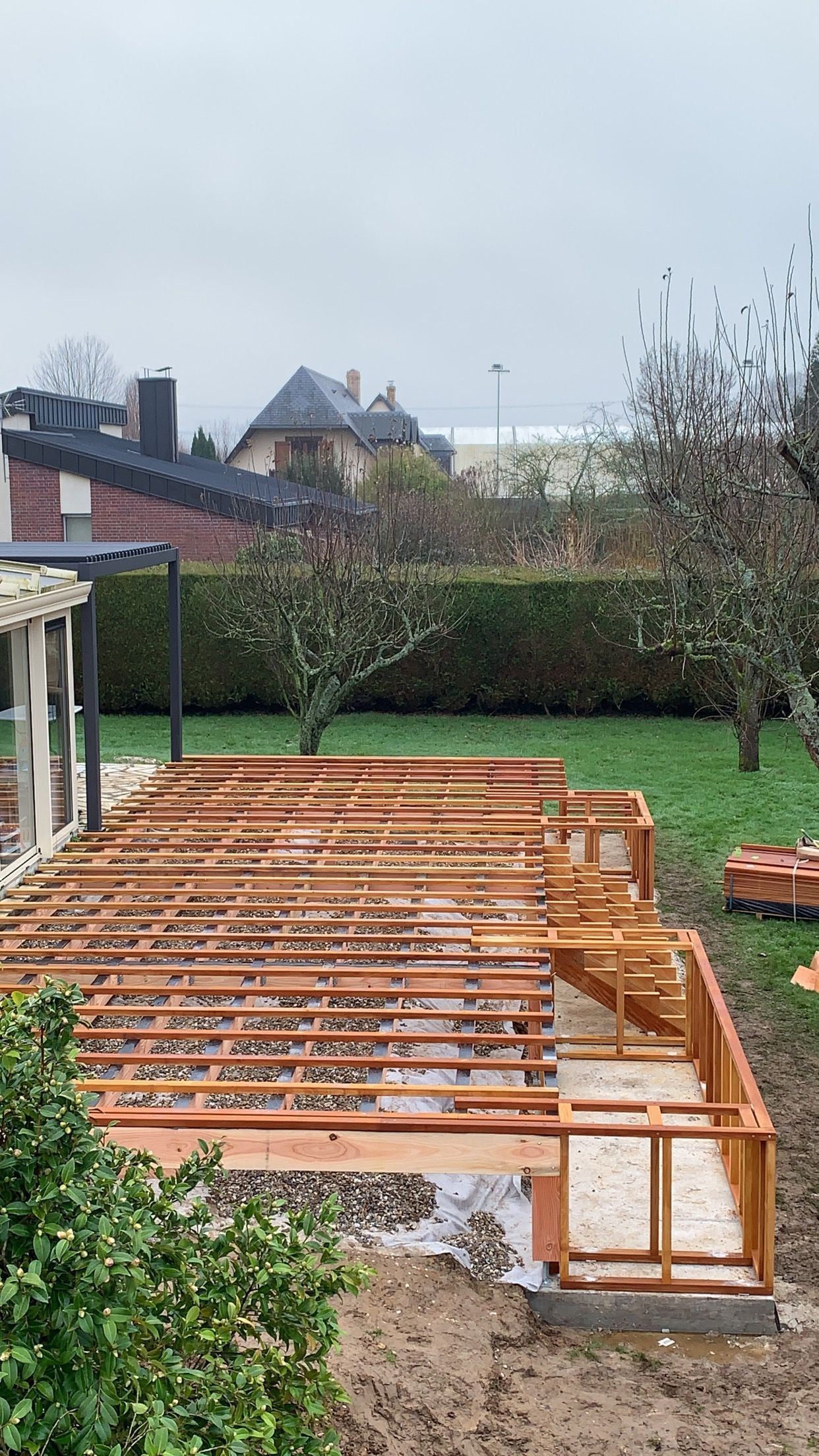 Construction d'une structure de terrasse en bois dans un jardin, entourée de verdure et sous un ciel couvert.