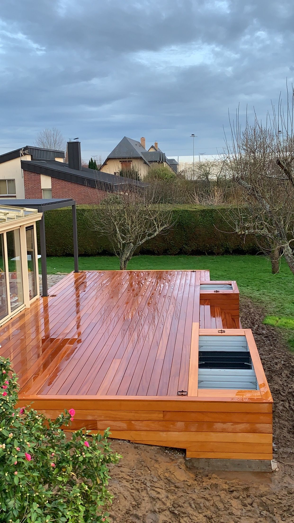 Terrasse en bois dans un jardin, mouillée par la pluie. Maison et végétation en arrière-plan sous un ciel couvert.